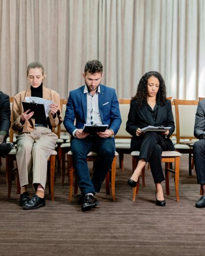 Five people wearing business attire, sitting on a row of chairs in a straight line, looking down at clipboards.