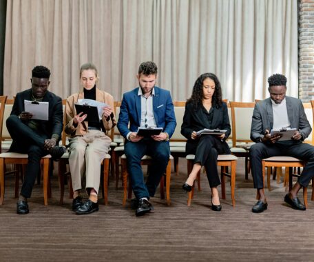 Five people wearing business attire, sitting on a row of chairs in a straight line, looking down at clipboards.